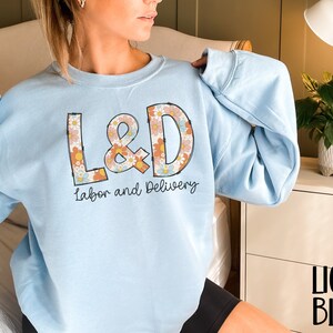 May include: Light blue sweatshirt with the text "L&D Labor and Delivery" in a retro floral design. The sweatshirt is a light blue color and has a crew neck. The text "LIGHT BLUE" is printed in black at the bottom right.
