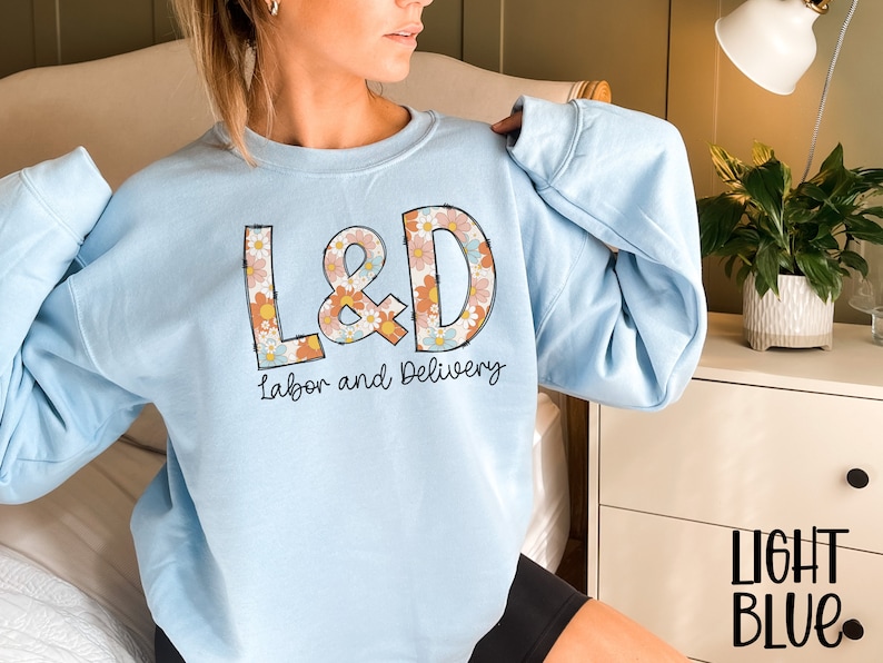 May include: Light blue sweatshirt with the text "L&D Labor and Delivery" in a retro floral design. The sweatshirt is a light blue color and has a crew neck. The text "LIGHT BLUE" is printed in black at the bottom right.