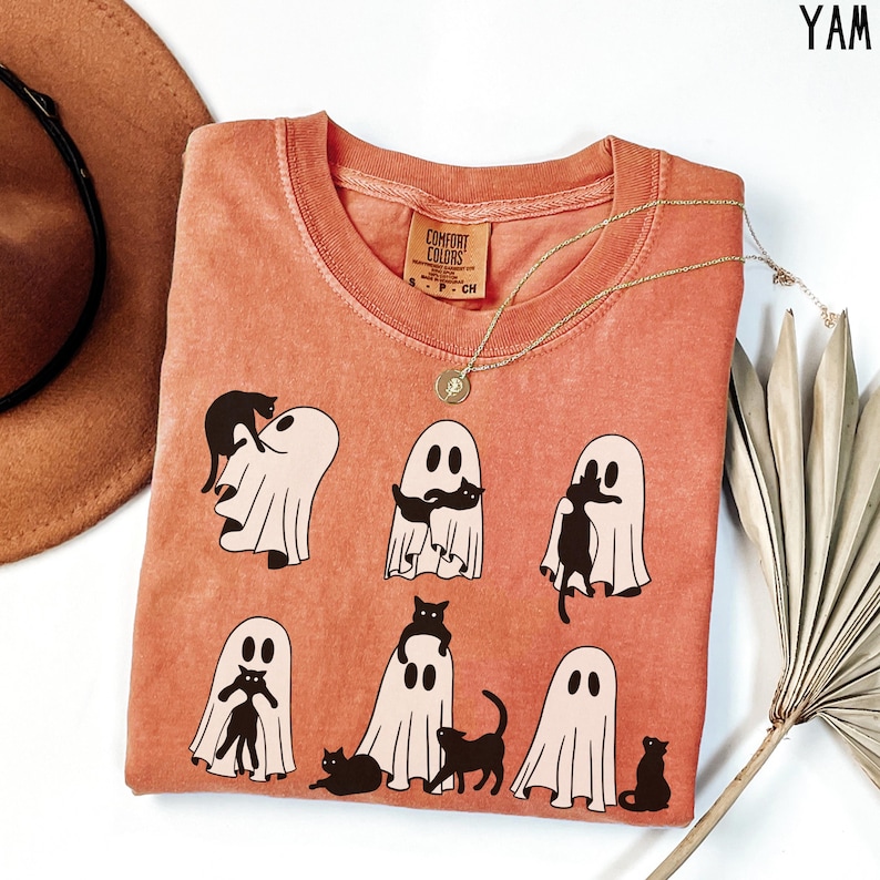 May include: A coral orange t-shirt with a graphic print of black cats and ghosts. The text "Comfort Colors" is on a white tag attached to the shirt.
