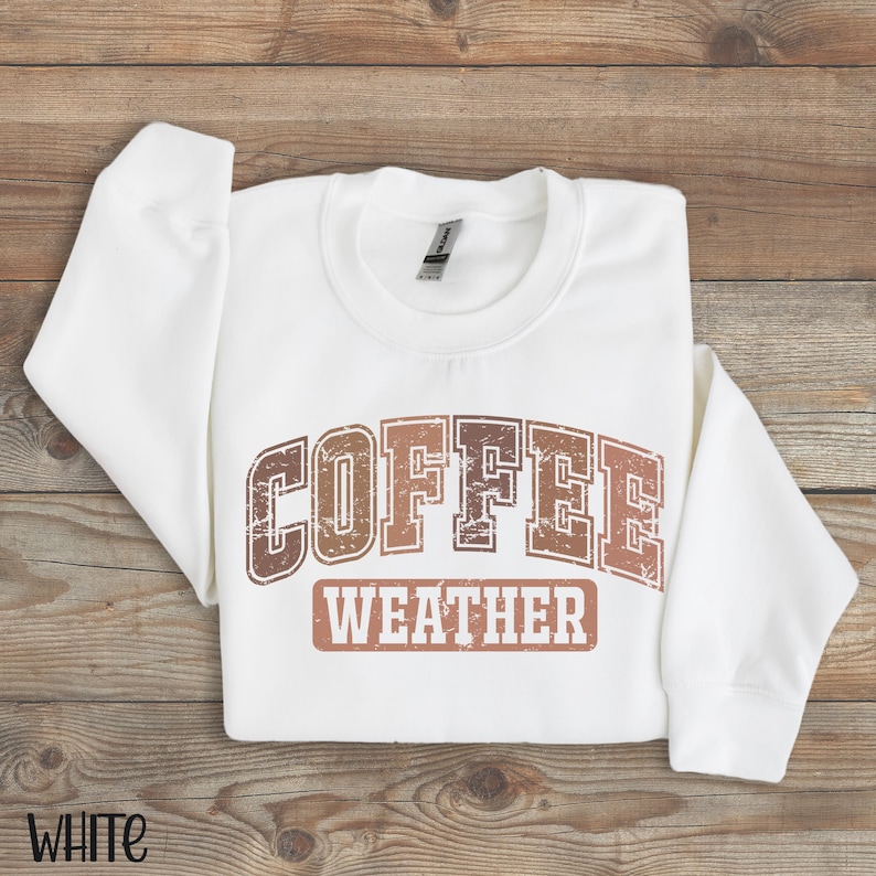 May include: White sweatshirt with the words "COFFEE WEATHER" in a brown, distressed font. The sweatshirt is folded on a wooden surface. The word "WHITE" is printed in the bottom left corner.