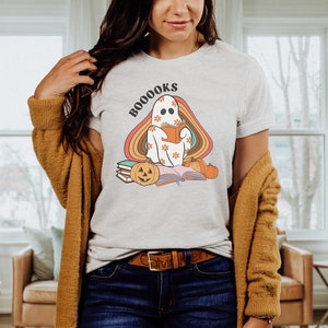 May include: A white t-shirt featuring a ghost reading a book with the text "BOO-OOOKS" in a retro font. The ghost is surrounded by a rainbow and there are pumpkins and books on the ground.