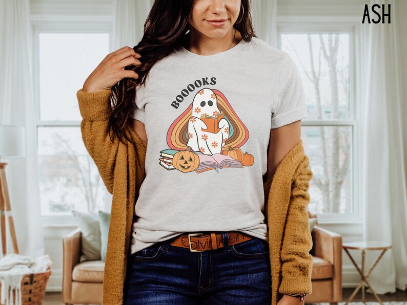 May include: A white t-shirt featuring a ghost reading a book with the text "BOO-OOOKS" in a retro font. The ghost is surrounded by a rainbow and there are pumpkins and books on the ground.