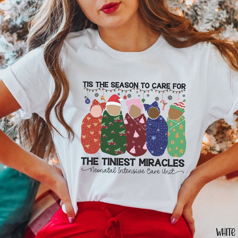 K&ouml;nnte beinhalten: Wei&szlig;es T-Shirt mit einem weihnachtlichen Design, das f&uuml;nf Babys zeigt, die in Decken mit verschiedenen Weihnachtsmustern gewickelt sind. Der Text lautet "Tis the season to care for the tiniest miracles Neonatal Intensive Care Unit".