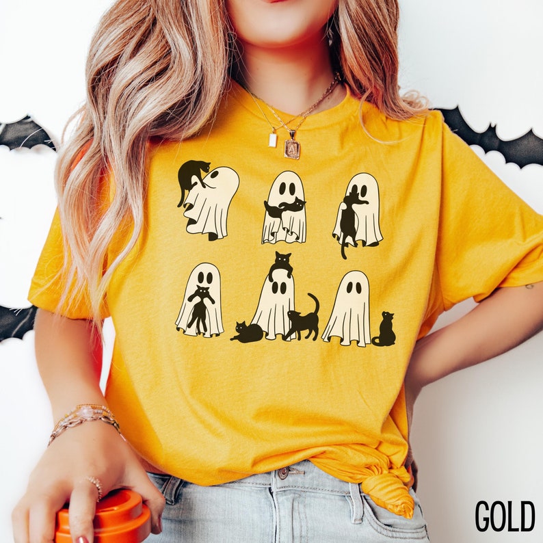 Puede incluir: Una camiseta amarilla con un gr&aacute;fico de gatos negros dentro de fantasmas blancos. El texto "GOLD" es visible en la esquina inferior derecha de la imagen.