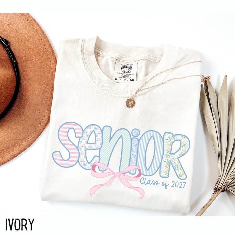 May include: Ivory-colored Comfort Colors t-shirt with the word "Senior" in pastel blue and pink patterns, a pink bow, and "Class of 2027" text. A gold necklace with a pendant and a brown hat are also visible.