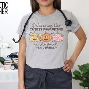 May include: Athletic heather gray t-shirt with the text "Delivering the cutest pumpkins in the patch L&D Nurse" and three cartoon pumpkins. The shirt is worn with gray scrubs and a stethoscope.