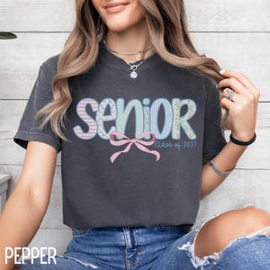 May include: A dark gray t-shirt with the word "SENIOR" in a colorful, patterned design. The word "SENIOR" is in a mix of pink, blue, and green patterns, with a pink bow below and "Class of 2027" printed below the bow.