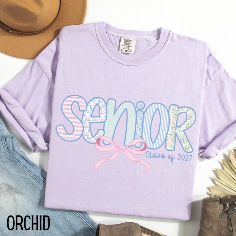 May include: A light purple t-shirt with the word "SENIOR" in a decorative font, featuring stripes, floral patterns, and a pink bow. The text "Class of 2027" is below the bow. The word "ORCHID" is at the bottom left.