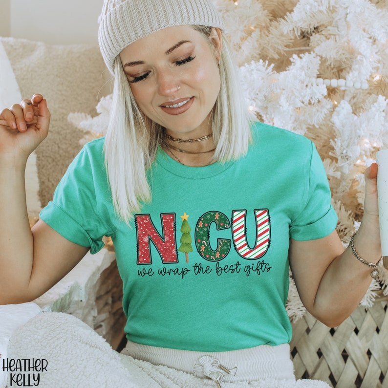 Puede incluir: Una camiseta verde turquesa con un dise&ntilde;o navide&ntilde;o que dice "NICU we wrap the best gifts". Las letras est&aacute;n decoradas con bastones de caramelo, hombres de jengibre y &aacute;rboles de Navidad.