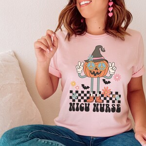 Puede incluir: Camiseta rosa con un gr&aacute;fico de una calabaza sonriente que lleva un sombrero de bruja y gafas de sol. La calabaza est&aacute; haciendo el signo de la paz y de pie sobre un patr&oacute;n a cuadros. El texto "NICU NURSE" est&aacute; impreso debajo de la calabaza.