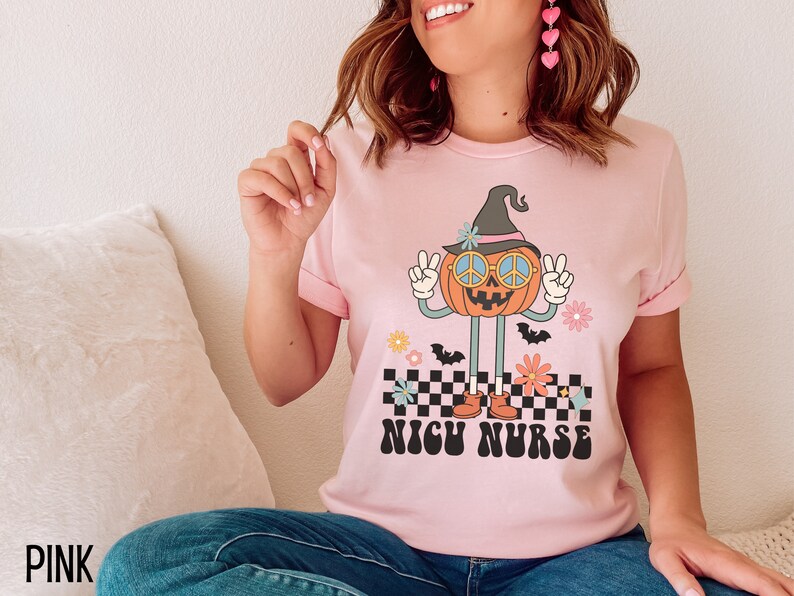 Puede incluir: Camiseta rosa con un gr&aacute;fico de una calabaza sonriente que lleva un sombrero de bruja y gafas de sol. La calabaza est&aacute; haciendo el signo de la paz y de pie sobre un patr&oacute;n a cuadros. El texto "NICU NURSE" est&aacute; impreso debajo de la calabaza.
