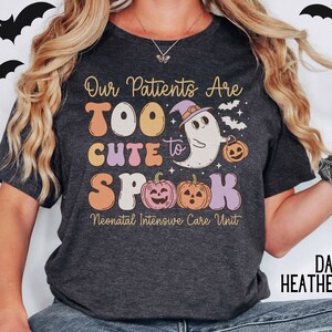 May include: Dark heather gray t-shirt with a cute ghost wearing a witch hat and holding a pumpkin. The text on the shirt reads "Our Patients Are Too Cute to Spook Neonatal Intensive Care Unit".
