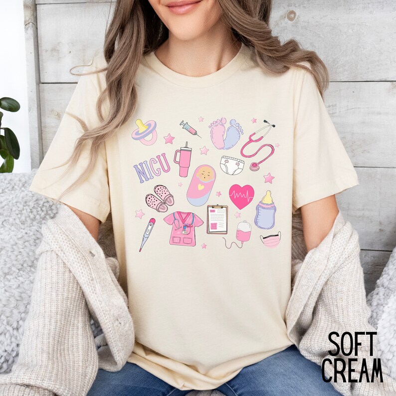 Puede incluir: Una camiseta de color crema con un dise&ntilde;o gr&aacute;fico rosa y azul que presenta varios s&iacute;mbolos m&eacute;dicos y la palabra "NICU".