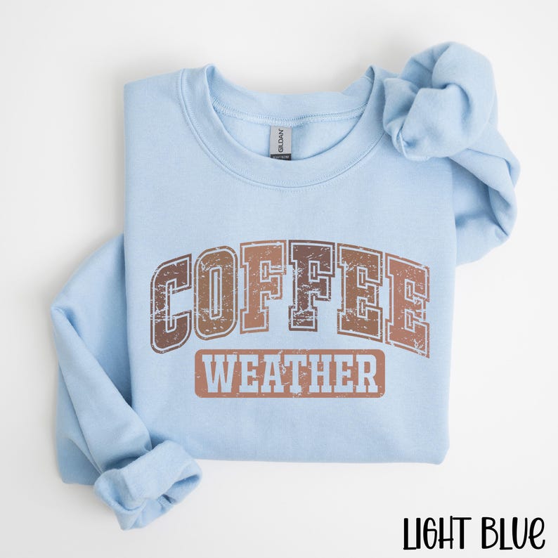 May include: A light blue sweatshirt with the words "COFFEE WEATHER" printed in brown and copper-colored letters. The sweatshirt is folded neatly, with the sleeves and collar visible. The text "LIGHT BLUE" is at the bottom right.