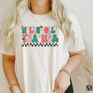 May include: A white t-shirt with the text "Holly Jolly Teacher" in colorful retro lettering. The text is surrounded by small green leaves and red and pink flowers.