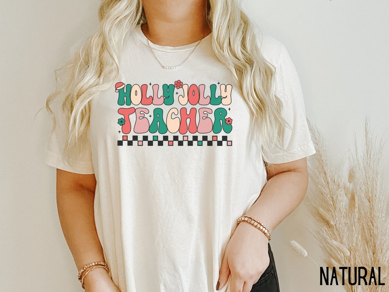 May include: A white t-shirt with the text "Holly Jolly Teacher" in colorful retro lettering. The text is surrounded by small green leaves and red and pink flowers.