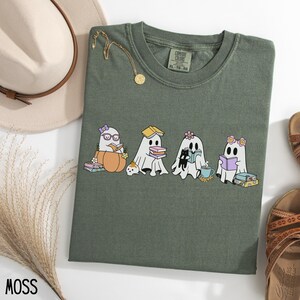 May include: Moss green t-shirt featuring a graphic of four cartoon ghosts reading books. One ghost is in a pumpkin, another has a book on its head, and a third is with a black cat. The shirt has a crew neck and short sleeves.