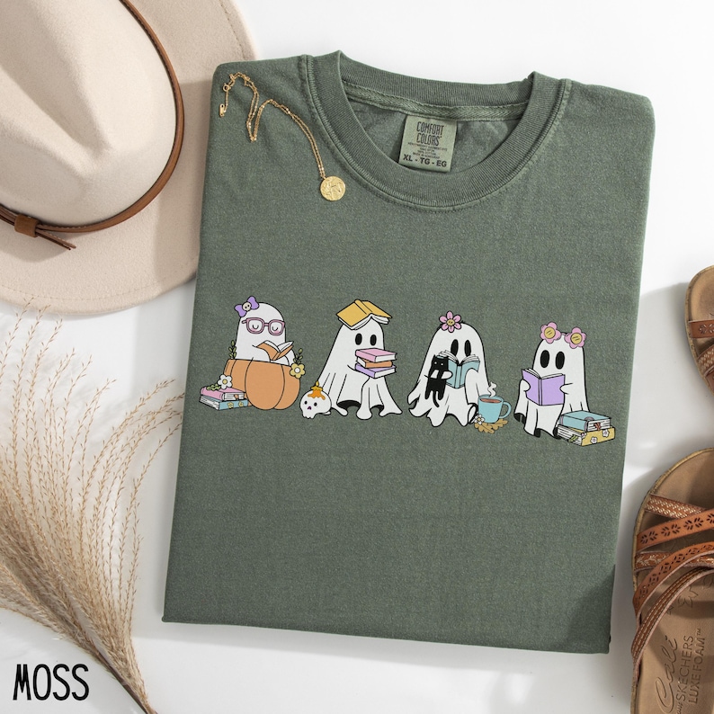 May include: Moss green t-shirt featuring a graphic of four cartoon ghosts reading books. One ghost is in a pumpkin, another has a book on its head, and a third is with a black cat. The shirt has a crew neck and short sleeves.