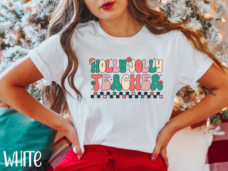 May include: White t-shirt with a retro-style graphic that says "Holly Jolly Teacher" in colorful letters with a red Santa hat, green and pink flowers, and a black and white checkered pattern.