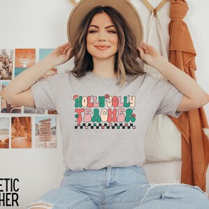 May include: A heather grey t-shirt with a retro-style graphic that says "Holly Jolly Teacher" in colorful letters with a Santa hat on the "H".