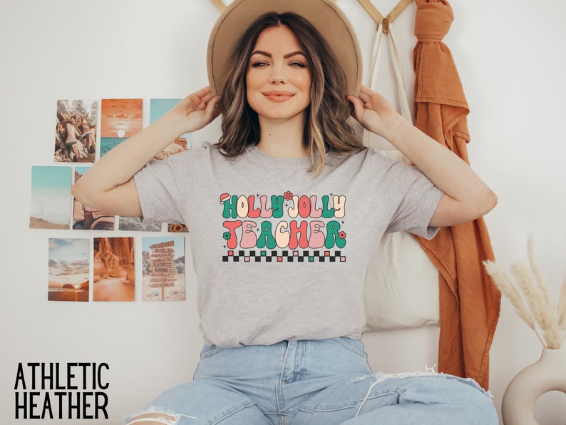 May include: A heather grey t-shirt with a retro-style graphic that says "Holly Jolly Teacher" in colorful letters with a Santa hat on the "H".