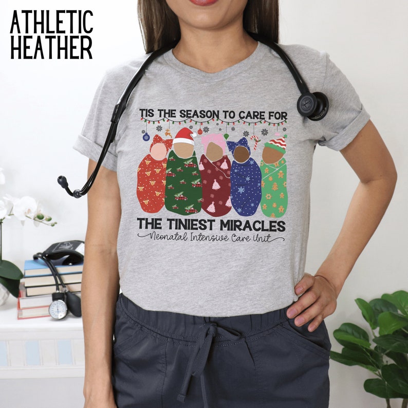 K&ouml;nnte beinhalten: Ein graues T-Shirt mit dem Text "Tis the season to care for the tiniest miracles - Neonatal Intensive Care Unit" und einer Grafik von f&uuml;nf Babys, die in Weihnachts-Swaddles gewickelt sind.