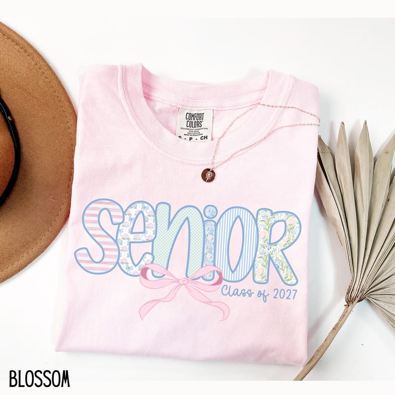 May include: A light pink t-shirt with the word "Senior" in a decorative font, featuring floral and striped patterns. A pink bow is below the word, with "Class of 2027" underneath. A gold necklace and a brown hat are also in the image.
