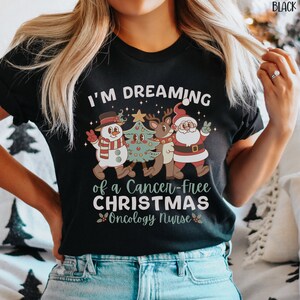 May include: Black t-shirt with a Christmas-themed design. The text reads "I'm dreaming of a cancer-free Christmas Oncology Nurse". The design features a snowman, a reindeer, and Santa Claus.
