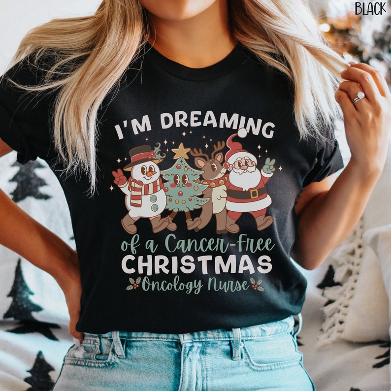 May include: Black t-shirt with a Christmas-themed design. The text reads "I'm dreaming of a cancer-free Christmas Oncology Nurse". The design features a snowman, a reindeer, and Santa Claus.