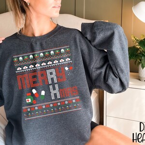 K&ouml;nnte beinhalten: Dunkelgrauer Sweatshirt mit einem weihnachtlichen Design mit dem Text "Merry & Mugs" und einem Muster aus roten und gr&uuml;nen Pillen, wei&szlig;en Krankenschwesterh&uuml;ten und roten und gr&uuml;nen Geschenkboxen.