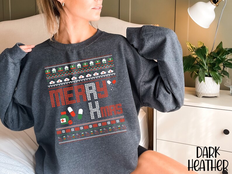 K&ouml;nnte beinhalten: Dunkelgrauer Sweatshirt mit einem weihnachtlichen Design mit dem Text "Merry & Mugs" und einem Muster aus roten und gr&uuml;nen Pillen, wei&szlig;en Krankenschwesterh&uuml;ten und roten und gr&uuml;nen Geschenkboxen.