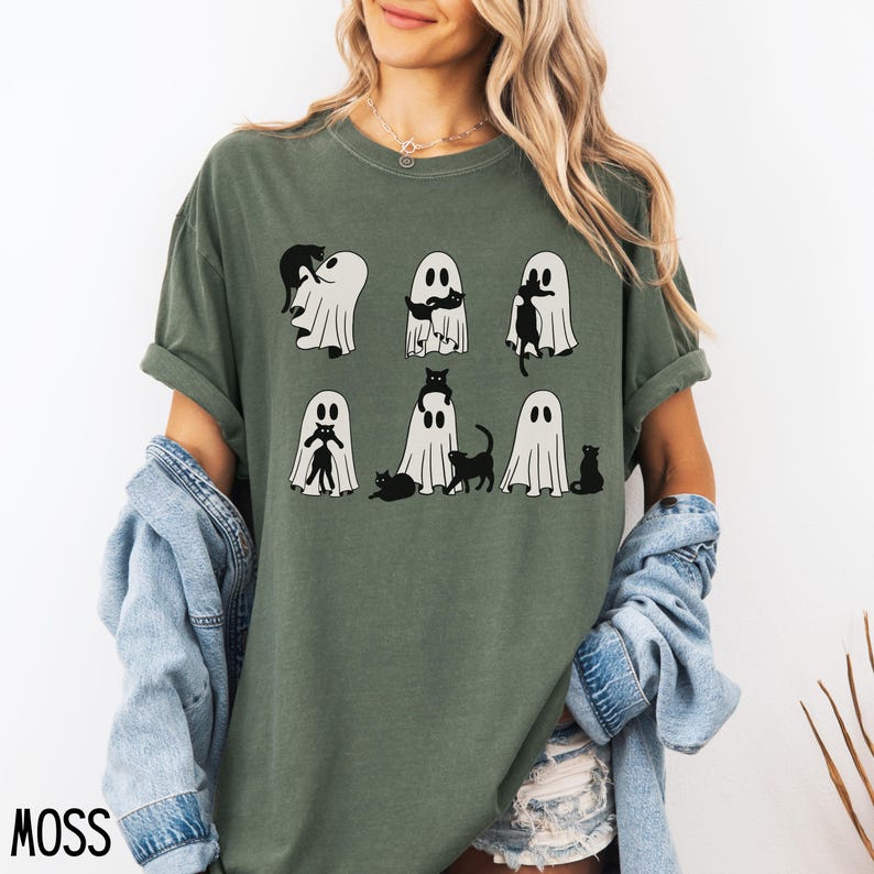 May include: A green t-shirt with a graphic of black cats and ghosts. The ghosts are holding the cats. The text on the shirt says "MOSS".