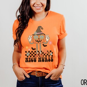 Puede incluir: Camiseta naranja con un gr&aacute;fico de una calabaza sonriente que lleva un sombrero de bruja y gafas de sol con el signo de la paz. La calabaza est&aacute; de pie sobre un patr&oacute;n a cuadros con el texto "NICU NURSE" debajo.