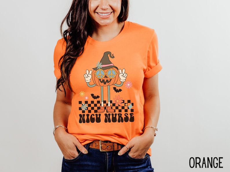Puede incluir: Camiseta naranja con un gr&aacute;fico de una calabaza sonriente que lleva un sombrero de bruja y gafas de sol con el signo de la paz. La calabaza est&aacute; de pie sobre un patr&oacute;n a cuadros con el texto "NICU NURSE" debajo.