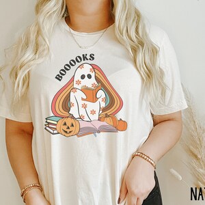 May include: A white t-shirt featuring a ghost reading a book with the text "BOO-OOOKS" in rainbow colours. The ghost is wearing a floral pattern and there are pumpkins and books around it.