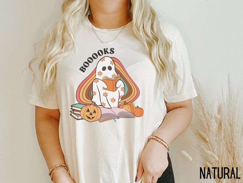 May include: A white t-shirt featuring a ghost reading a book with the text "BOO-OOOKS" in rainbow colours. The ghost is wearing a floral pattern and there are pumpkins and books around it.