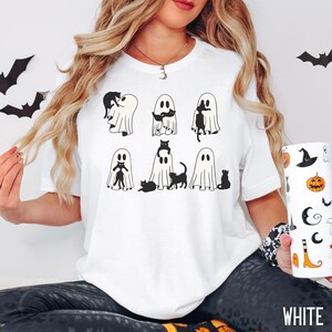 Puede incluir: Camiseta blanca con un gr&aacute;fico de seis fantasmas, cada uno con un gato negro dentro. Los fantasmas est&aacute;n en diferentes poses, algunos con el gato asom&aacute;ndose, otros con el gato abrazando al fantasma y otros con el gato sentado en la cabeza del fantasma. El texto "WHITE" est&aacute; en la parte inferior de la imagen.
