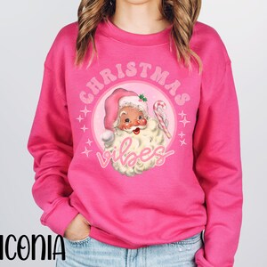 May include: Pink Christmas sweatshirt with a retro illustration of Santa Claus wearing a pink hat and holding a candy cane. The text "Christmas Vibes" is written in pink letters around the illustration.