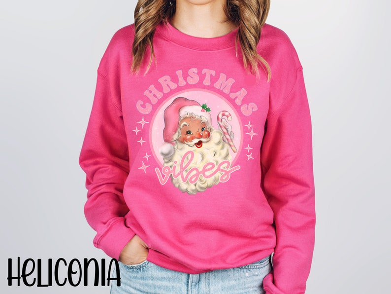 May include: Pink Christmas sweatshirt with a retro illustration of Santa Claus wearing a pink hat and holding a candy cane. The text "Christmas Vibes" is written in pink letters around the illustration.