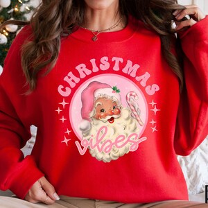 May include: Red Christmas sweatshirt with a pink circle graphic featuring a cartoon Santa Claus with the text "Christmas Vibes".