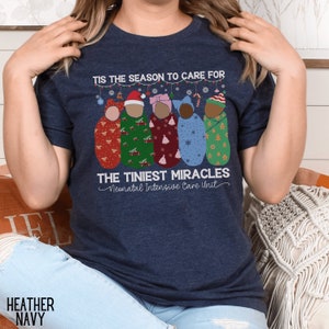 K&ouml;nnte beinhalten: Ein marineblaues T-Shirt mit dem Text "Tis the season to care for the tiniest miracles Neonatal Intensive Care Unit". Das Design zeigt f&uuml;nf gewickelte Babys in Weihnachtsoutfits.