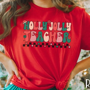 May include: Red t-shirt with a retro-style graphic that says "Holly Jolly Teacher" in green, pink, and white.