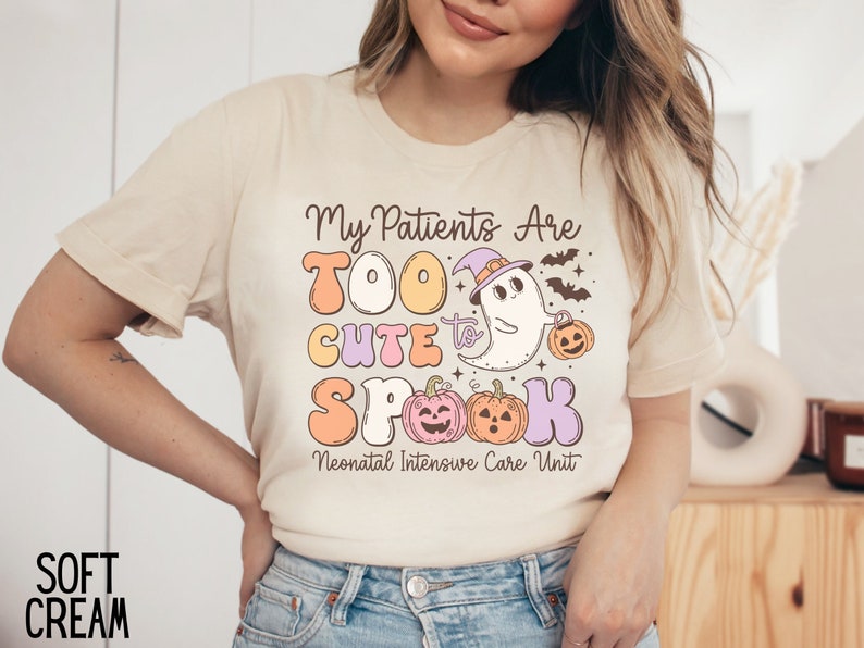 May include: A cream colored t-shirt with the text "My Patients Are Too Cute to Spook" in colorful letters. A cartoon ghost wearing a witch hat and holding a pumpkin is above the text. The text "Neonatal Intensive Care Unit" is below the graphic.