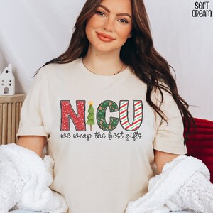 Puede incluir: Una camiseta de color crema con un dise&ntilde;o navide&ntilde;o que dice "NICU we wrap the best gifts". Las letras est&aacute;n decoradas con patrones rojos, verdes y blancos.