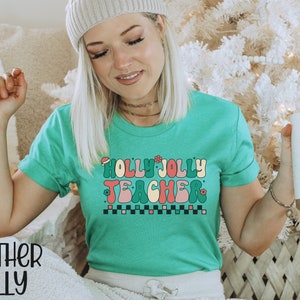 May include: A green t-shirt with the text "Holly Jolly Teacher" in a retro font with a red Santa hat, green holly leaves, and pink flowers. The text is in a colorful, wavy font.