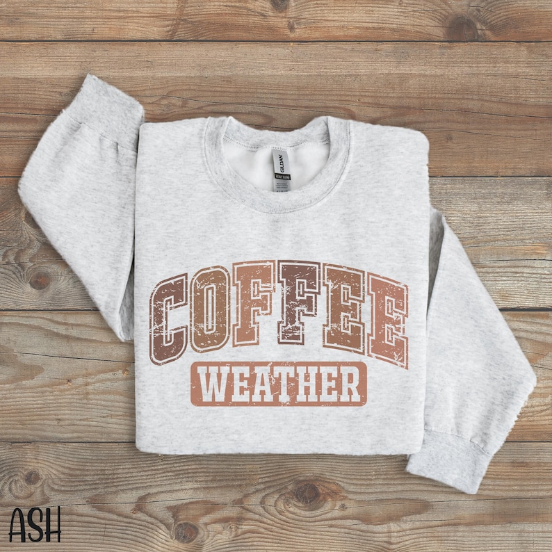 May include: A light gray sweatshirt with the words "COFFEE WEATHER" in a brown, distressed font. The sweatshirt has long sleeves and a crew neck. The text is in a collegiate style.