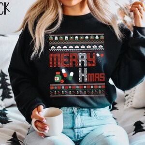 K&ouml;nnte beinhalten: Schwarzer Weihnachtspullover mit rotem und wei&szlig;em Muster und dem Text "Merry Xmas". Der Pullover zeigt verschiedene medizinische Symbole, darunter Pillen, ein Stethoskop und eine Flasche Medizin.