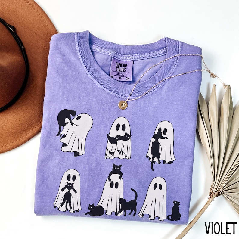 May include: A purple t-shirt with a graphic of black cats and ghosts. The text "Comfort Colors" is on the tag.