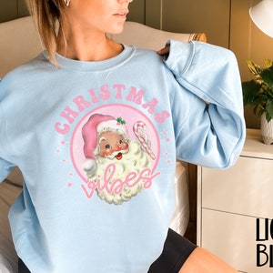 May include: Light blue sweatshirt with a pink Christmas-themed graphic featuring a cartoon Santa Claus with the text "Christmas Vibes".