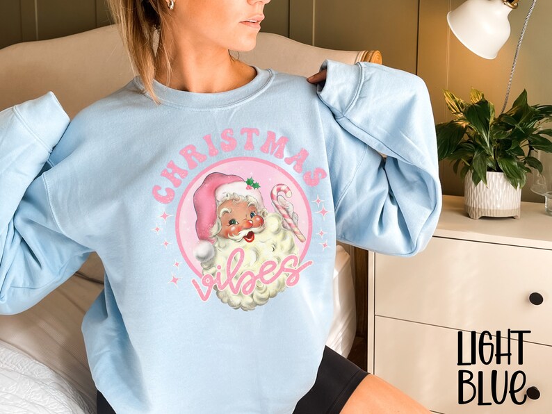 May include: Light blue sweatshirt with a pink Christmas-themed graphic featuring a cartoon Santa Claus with the text "Christmas Vibes".